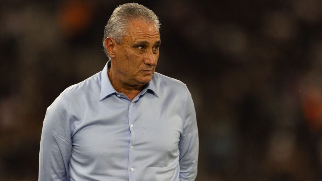 Homem de cabelos grisalhos em pé, usando camisa azul claro. Ele olha para o lado em um ambiente escuro.