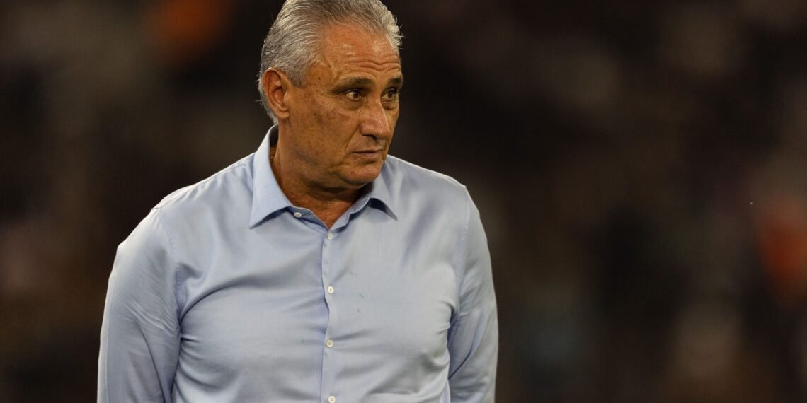 Homem de cabelos grisalhos em pé, usando camisa azul claro. Ele olha para o lado em um ambiente escuro.