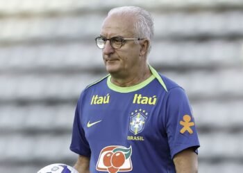 Homem com camisa do Brasil, segurando uma bola de futebol, em ambiente de treino. Campos desfocados ao fundo.