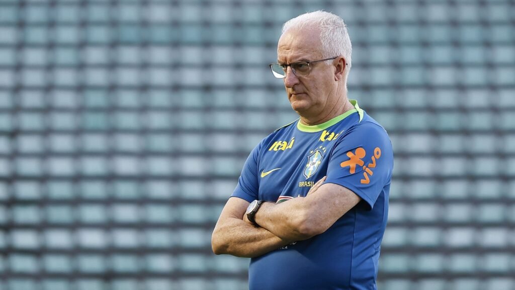 Técnico de futebol concentrado, com uniforme da seleção, observa treinamento em campo vazio com arquibancadas ao fundo.