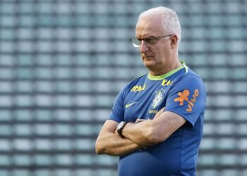 Técnico de futebol concentrado, com uniforme da seleção, observa treinamento em campo vazio com arquibancadas ao fundo.