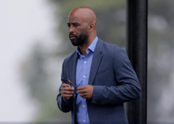 Corinthians planeja contratações seguras e sustentáveis 39 Homem de terno azul, segurando óculos, parece pensativo em ambiente ao ar livre. Fundo desfocado e verde.