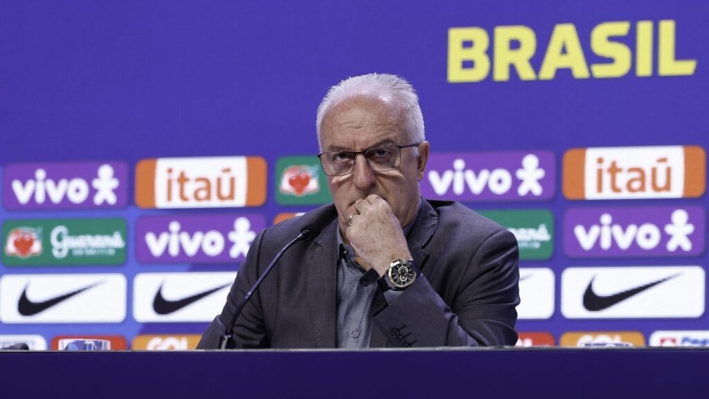 Homem em conferência de imprensa, pensativo, com logotipos ao fundo, destacando marcas e associações ao Brasil.