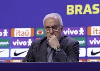 Corinthians próxima de fechar com Dorival em nova reunião 40 Homem em conferência de imprensa, pensativo, com logotipos ao fundo, destacando marcas e associações ao Brasil.
