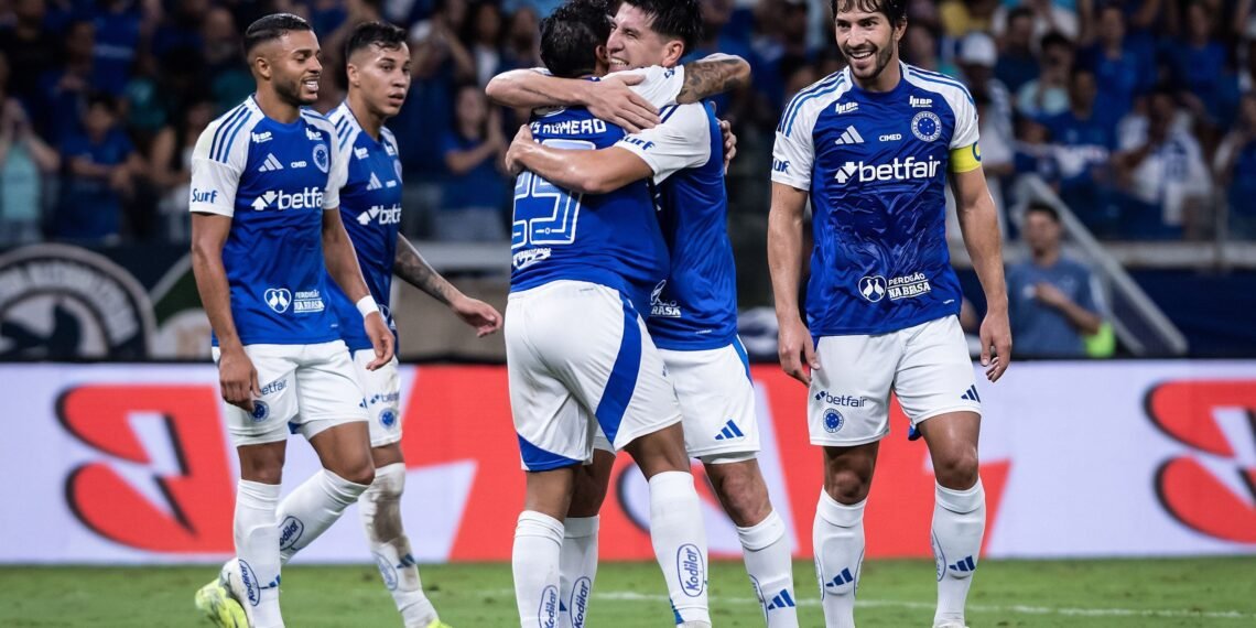Jogadores de futebol do time Cruzeiro comemoram no campo, usando uniformes azuis e brancos, após um gol em partida.