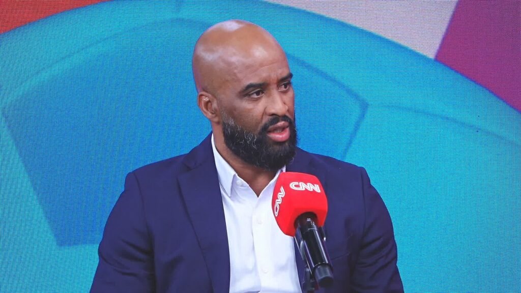 Homem de barba vestindo terno escuro dá entrevista ao vivo na TV, segurando microfone com logotipo da CNN.