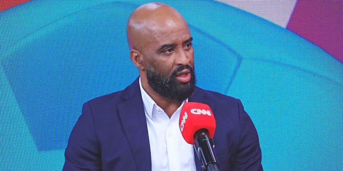 Homem de barba vestindo terno escuro dá entrevista ao vivo na TV, segurando microfone com logotipo da CNN.