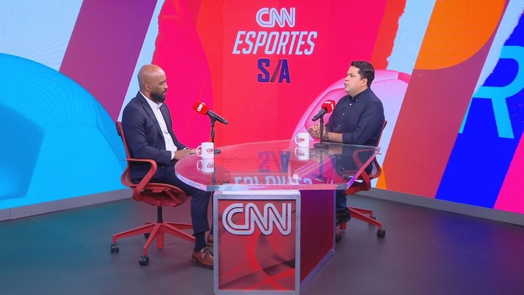 Dois homens conversam em estúdio moderno da CNN Esportes S/A, sentados frente a frente em uma mesa com microfones e canecas.