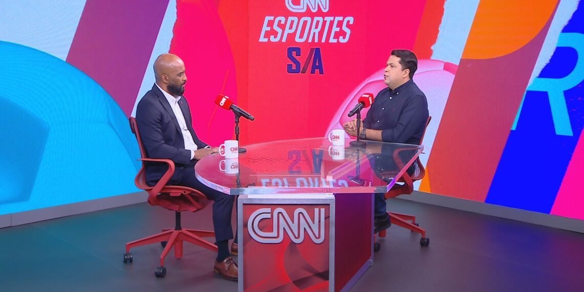 Dois homens conversam em estúdio moderno da CNN Esportes S/A, sentados frente a frente em uma mesa com microfones e canecas.