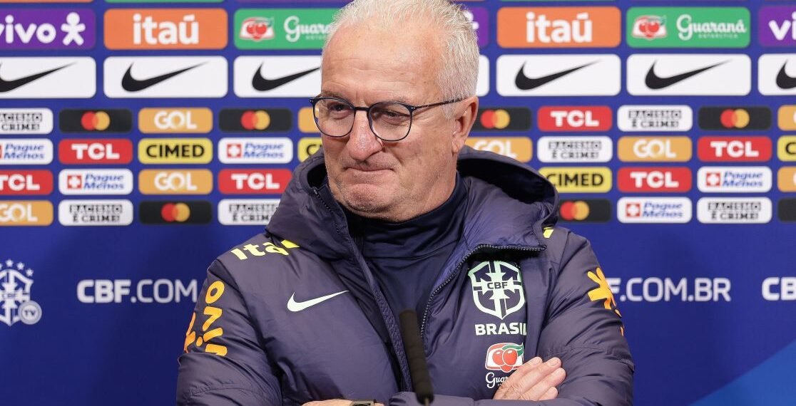 Treinador da seleção brasileira em coletiva de imprensa, usando agasalho da CBF, frente a painel de patrocinadores.