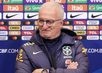 Treinador da seleção brasileira em coletiva de imprensa, usando agasalho da CBF, frente a painel de patrocinadores.