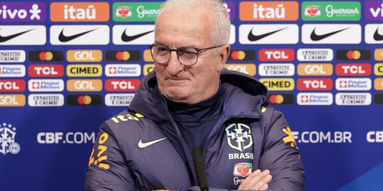 Treinador da seleção brasileira em coletiva de imprensa, usando agasalho da CBF, frente a painel de patrocinadores.