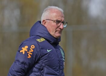 Treinador experiente observa treino usando jaqueta esportiva azul com logotipos de patrocinadores ao ar livre.