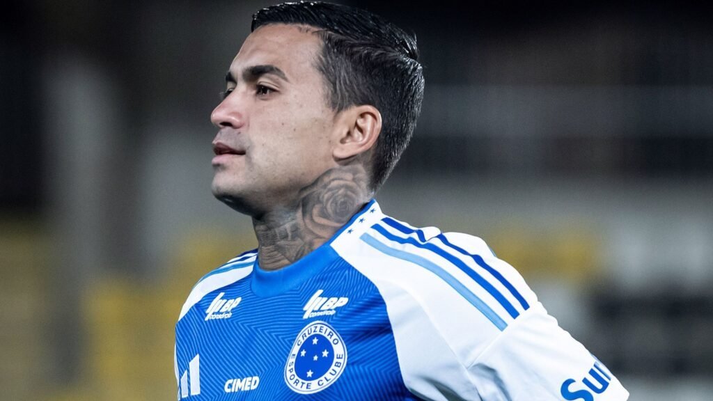 Jogador de futebol do Cruzeiro em campo, concentrado, veste uniforme azul com detalhes brancos, exibindo tatuagem no pescoço.