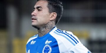 Jogador de futebol do Cruzeiro em campo, concentrado, veste uniforme azul com detalhes brancos, exibindo tatuagem no pescoço.