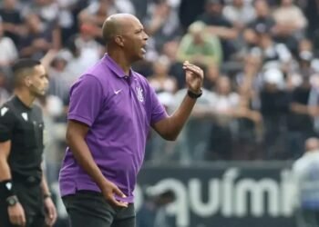 Técnico orienta jogadores à beira do campo durante jogo de futebol, vestido com camisa roxa, com público ao fundo.