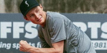 Jovem com boné preto e camiseta cinza comemora ao ar livre, exibindo expressão sorridente diante de um fundo esportivo.