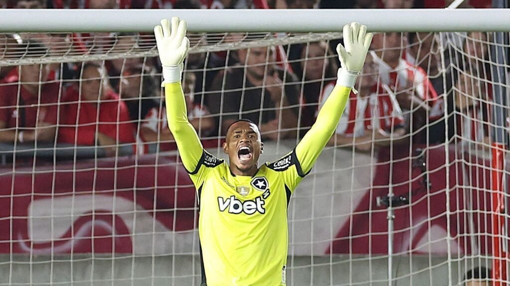 Goleiro de uniforme amarelo com mãos levantadas faz defesa em jogo de futebol, cercado por torcida ao fundo.