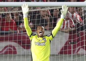 Goleiro de uniforme amarelo com mãos levantadas faz defesa em jogo de futebol, cercado por torcida ao fundo.