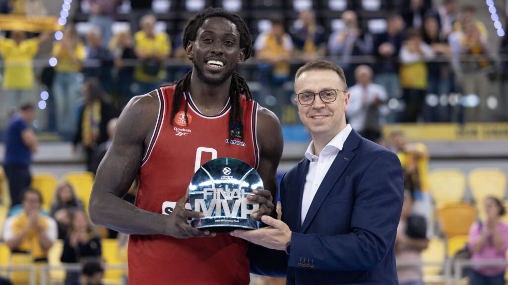 Jogador de basquete recebe troféu de MVP das finais, ao lado de oficial sorridente, em quadra com torcida ao fundo.