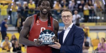 Jogador de basquete recebe troféu de MVP das finais, ao lado de oficial sorridente, em quadra com torcida ao fundo.