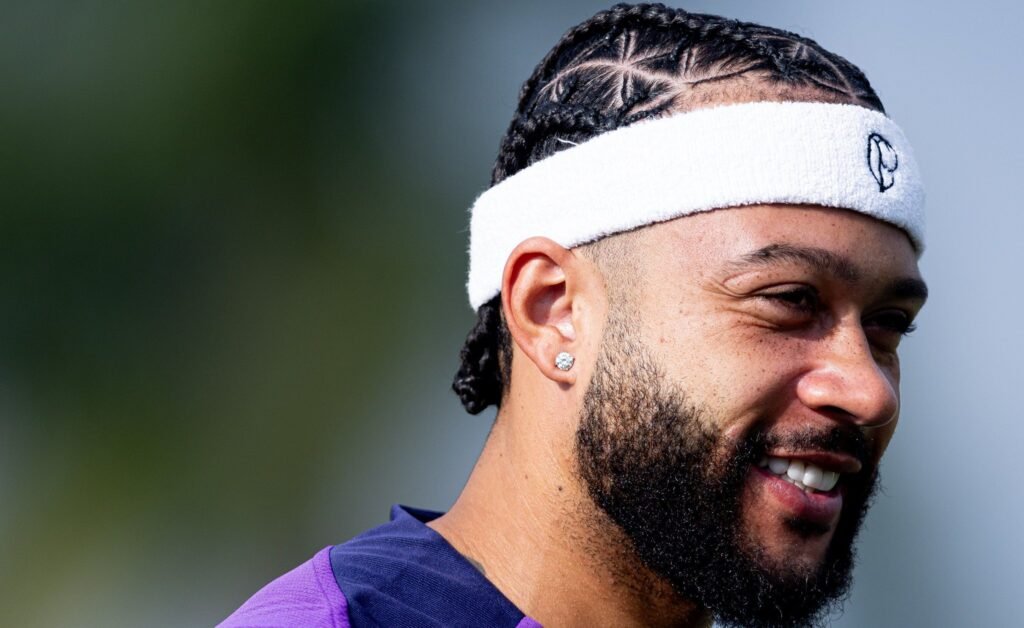 Homem com cabelo trançado e faixa esportiva branca sorri enquanto olha para a frente, em ambiente externo durante o dia.