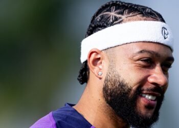 Homem com cabelo trançado e faixa esportiva branca sorri enquanto olha para a frente, em ambiente externo durante o dia.