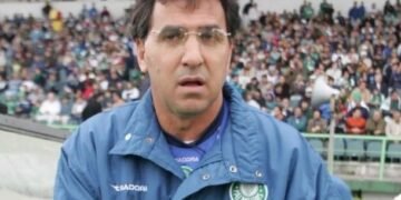 Homem usando jaqueta do Palmeiras em estádio lotado, cercado por torcida, aparenta estar concentrado no ambiente esportivo.
