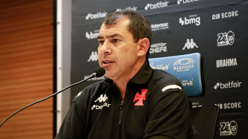Homem em coletiva de imprensa, vestindo camisa do Vasco, sentado em frente a microfone. Painel com logotipos ao fundo.