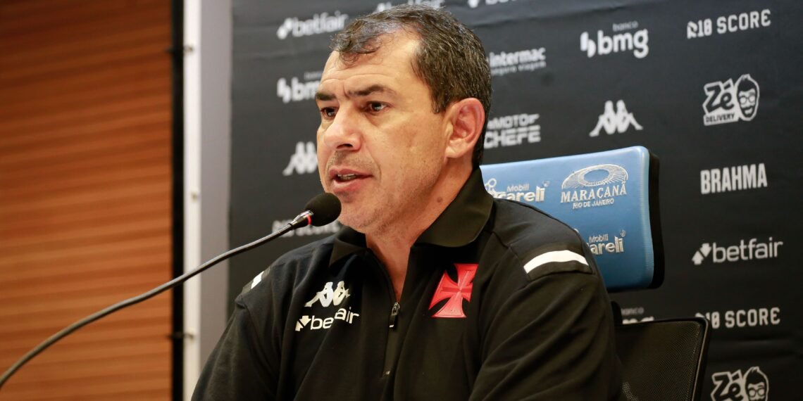 Homem em coletiva de imprensa, vestindo camisa do Vasco, sentado em frente a microfone. Painel com logotipos ao fundo.