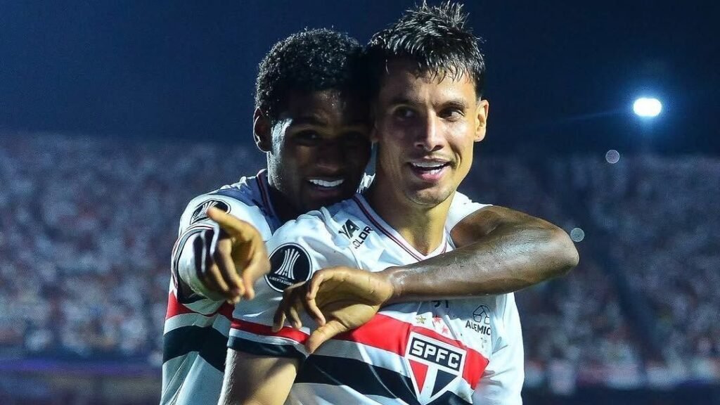 Jogadores do São Paulo comemoram gol em campo, abraçando-se e sorrindo, vestindo uniforme branco com escudo do clube.
