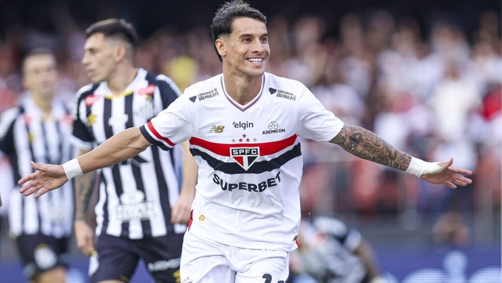 Jogador do São Paulo celebra gol em campo, vestindo uniforme branco com detalhes em preto e vermelho. Torcida ao fundo.