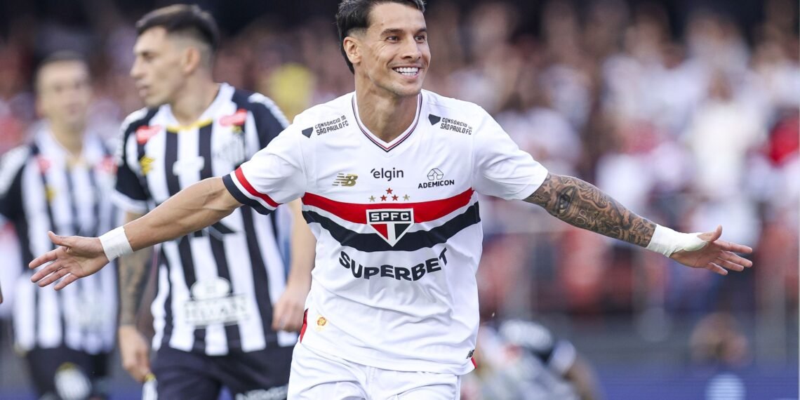Jogador do São Paulo celebra gol em campo, vestindo uniforme branco com detalhes em preto e vermelho. Torcida ao fundo.