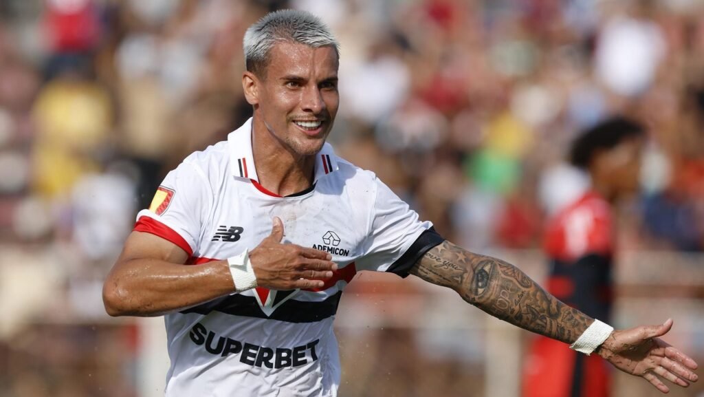 Jogador de futebol comemorando gol, exaltando emoção em campo, com uniforme branco e tatuagens visíveis no braço.