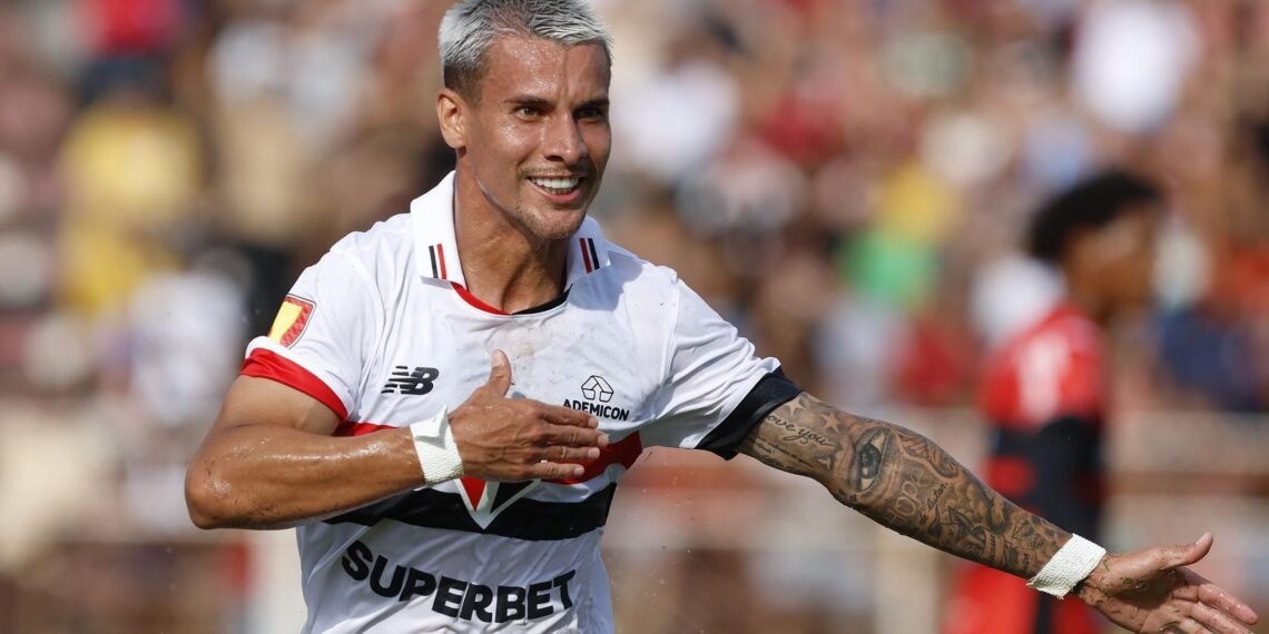 Jogador de futebol comemorando gol, exaltando emoção em campo, com uniforme branco e tatuagens visíveis no braço.