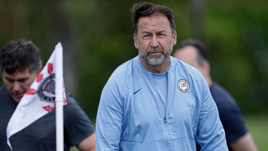 Técnico de futebol com jaqueta azul em campo, próximo a bandeira com símbolo do Corinthians, focado durante treino.