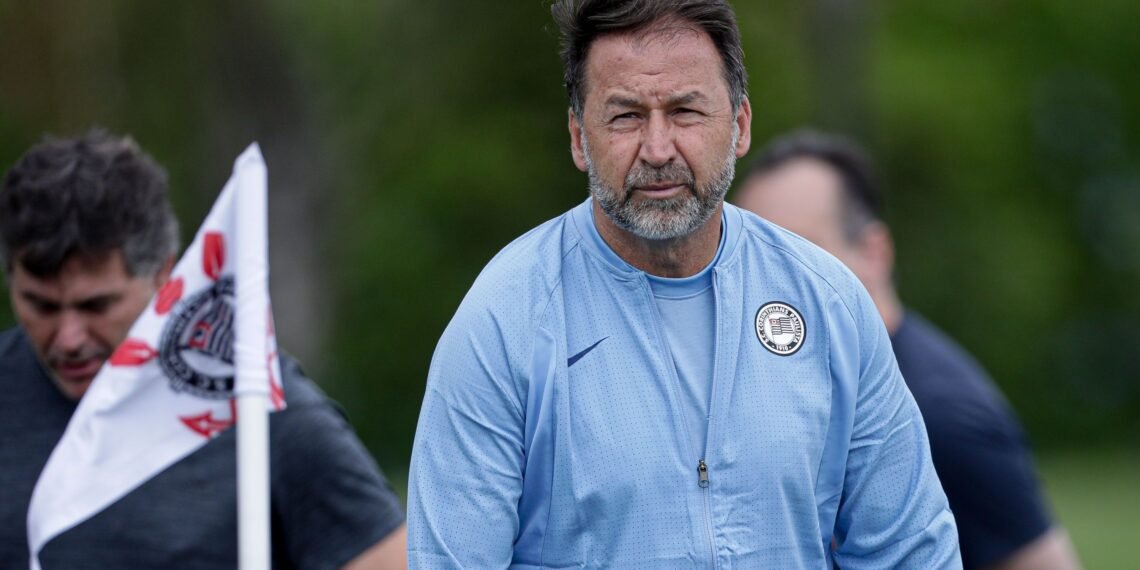 Técnico de futebol com jaqueta azul em campo, próximo a bandeira com símbolo do Corinthians, focado durante treino.