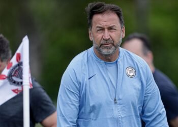 Fiel reúne-se com Augusto Melo: plano para a nação corinthiana 33 Técnico de futebol com jaqueta azul em campo, próximo a bandeira com símbolo do Corinthians, focado durante treino.