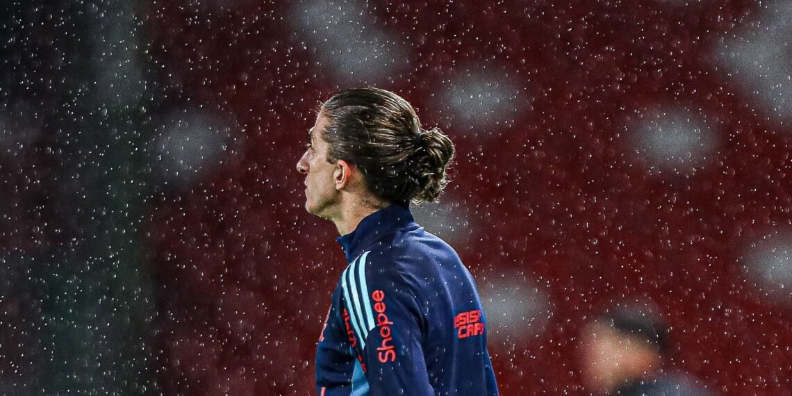 Jogador de futebol de perfil, com cabelo amarrado, em campo sob chuva, veste camisa azul com logotipo.