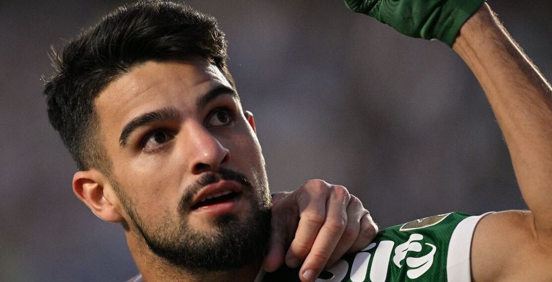 Jogador de futebol do Palmeiras celebra com expressão confiante, levantando o braço, em campo durante partida.