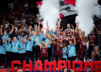 Equipe de basquete vence campeonato, comemorando com troféu em mãos, uniformes vermelhos e pretos, e fumaça branca.