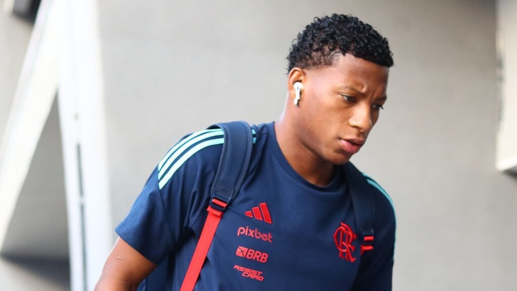 Pessoa com camiseta esportiva azul e vermelho, usando fones de ouvido, segura uma mochila e olha para baixo, caminhando.