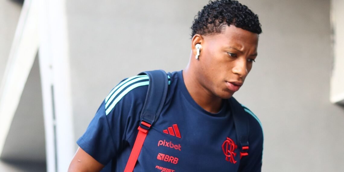 Pessoa com camiseta esportiva azul e vermelho, usando fones de ouvido, segura uma mochila e olha para baixo, caminhando.