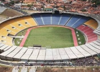 Vista aérea de um estádio de futebol com assentos coloridos, área de atletismo ao redor do campo e dezenas de pessoas reunidas na entrada.