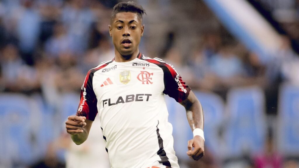 Jogador de futebol em ação veste o uniforme do Flamengo, com logotipo do clube visível, em um estádio lotado.