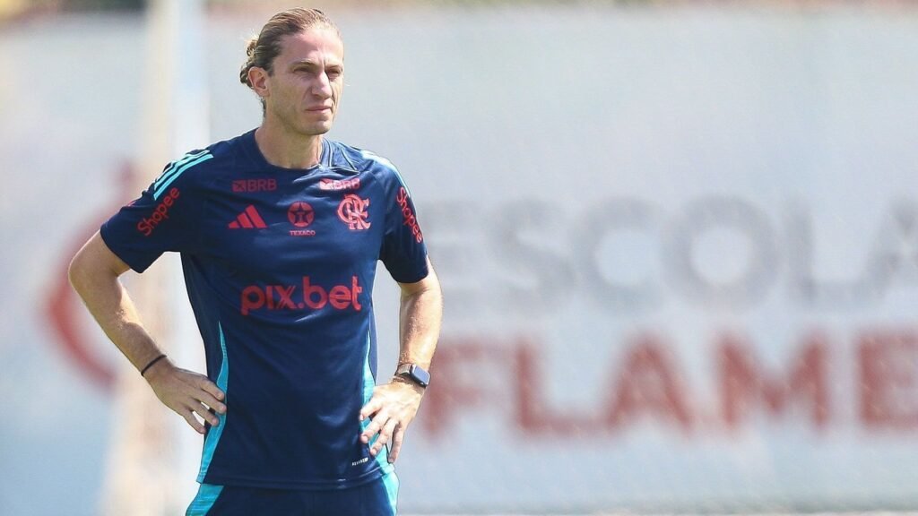 Treinador de futebol do Flamengo observa o campo, usando uniforme esportivo oficial do clube, mãos na cintura, expressão séria.