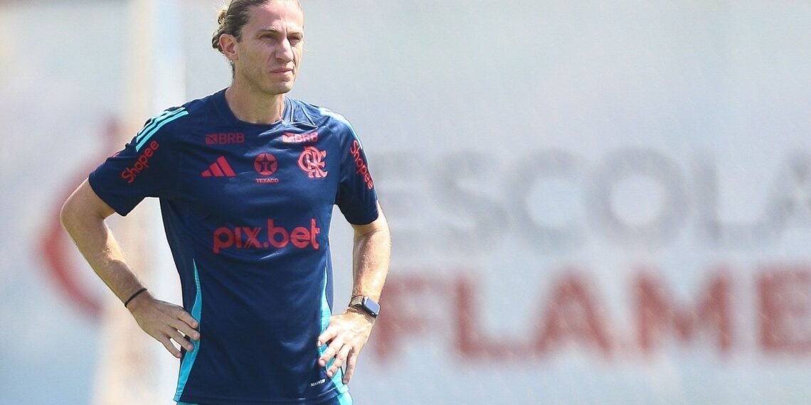 Treinador de futebol do Flamengo observa o campo, usando uniforme esportivo oficial do clube, mãos na cintura, expressão séria.