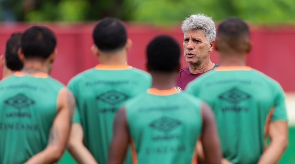 Treinador de futebol orienta jogadores durante treino; equipe em uniformes verdes presta atenção nas instruções no campo.