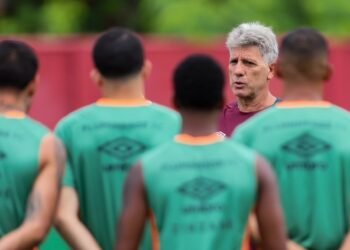 Treinador de futebol orienta jogadores durante treino; equipe em uniformes verdes presta atenção nas instruções no campo.