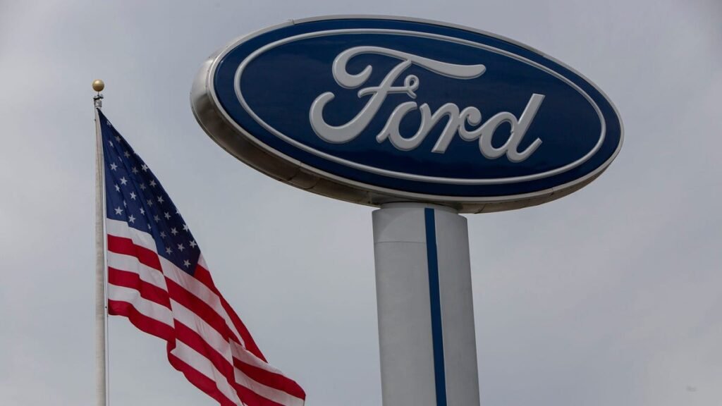 Placa da Ford ao lado de uma bandeira dos EUA em mastro alto, simbolizando a influência americana no setor automotivo.